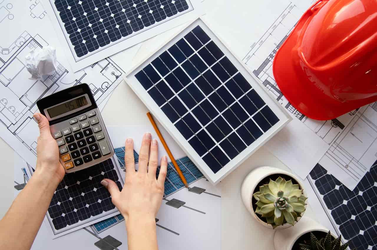 Mesa vista de cima com plantas de projeto de energia solar, um capacete de segurança vermelho e uma calculadora.