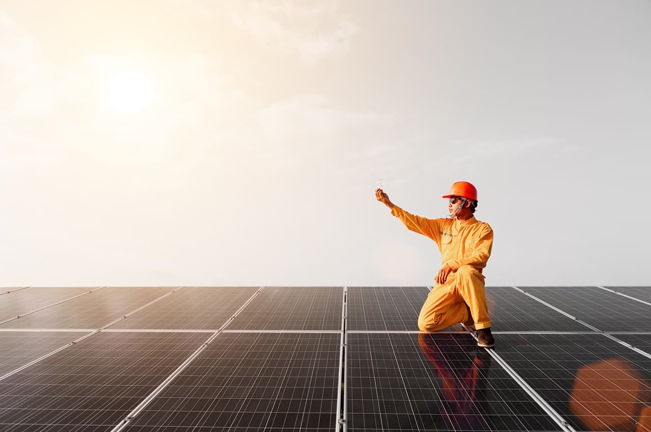 Homem ajoelhado em cima do painel solar, vestindo capacete e roupa de proteção laranja e segurando uma lâmpada acesa na mão direita.