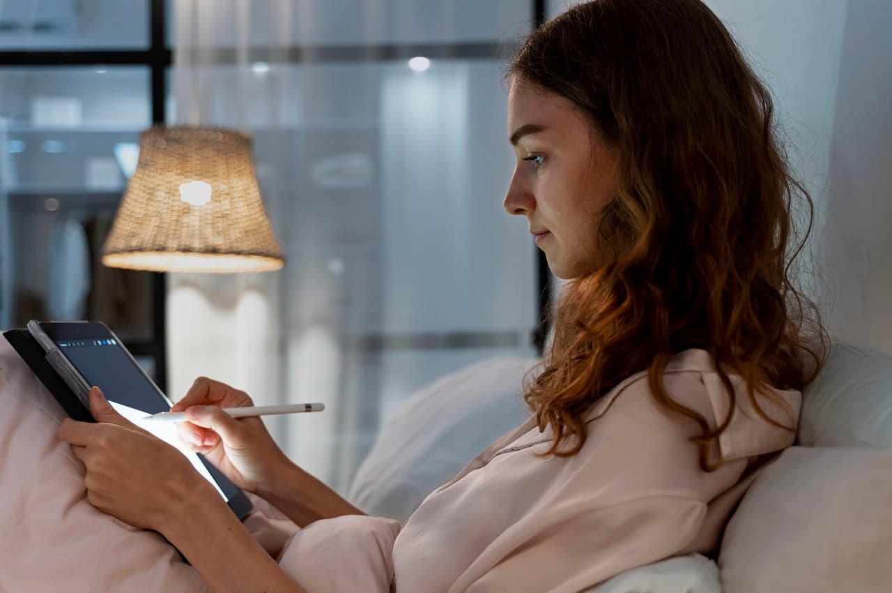 Mulher de pijama e expressão relaxada sentada na cama e com abajur ligado, enquanto digita no tablet.