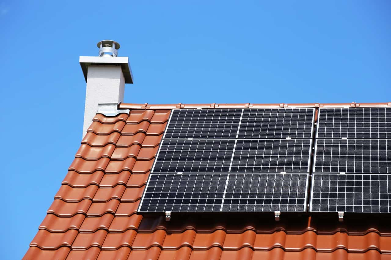 Painel solar instalado no telhado de uma casa com chaminé aparente. Ao fundo, tem um céu aberto e azul anil.