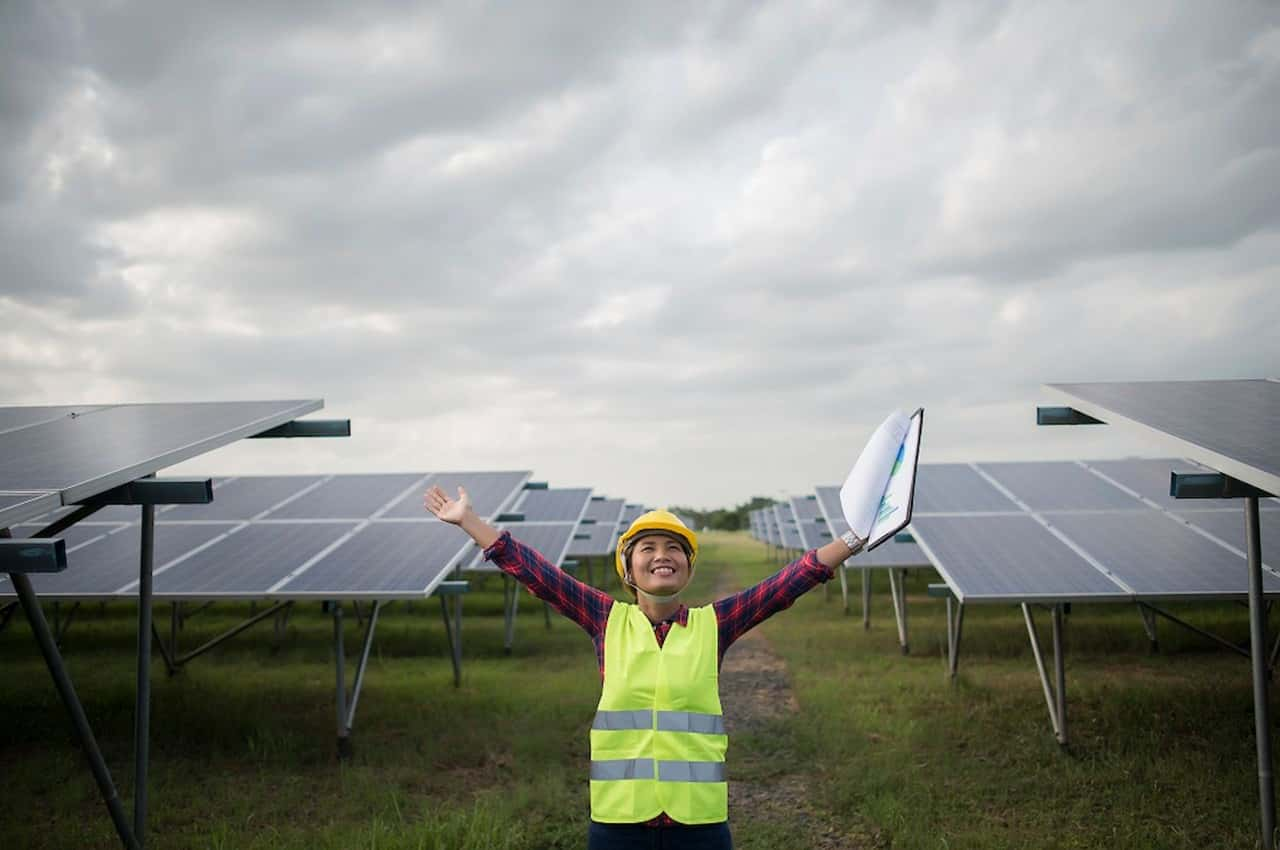 Mulher sorridente de braços abertos, vestindo EPI de segurança (capacete e colete) ao lado de painéis solares.