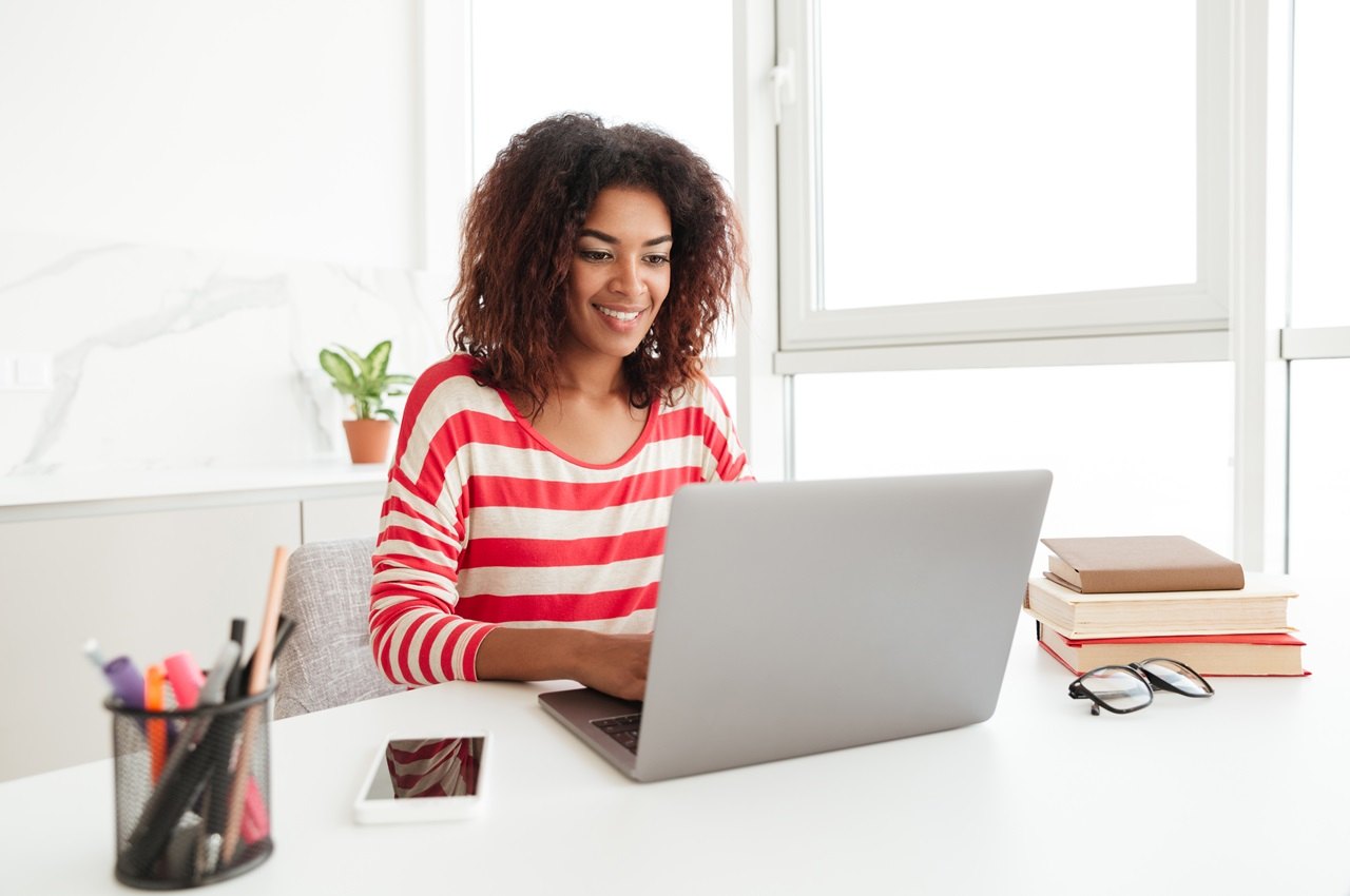 Mulher sorridente de blusa listrada vermelha e branca, digitando no computador que esta ao lado de livros e canetas.