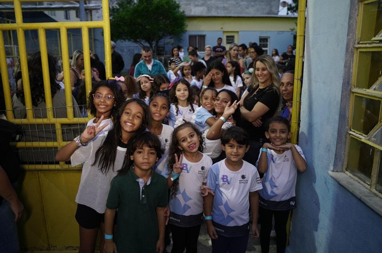 Grupo de crianças sorridentes no ambiente escolar vestindo camisetas azul e branca com a marca banco BV por conta da iniciativa BV Esportes.