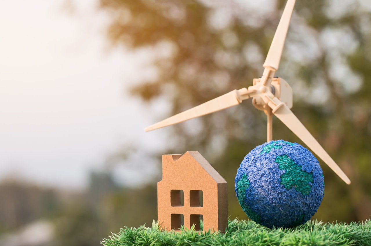 Pequena maquete de casa de madeira, ao lado de uma hélice de energia eólica, representando uma casa sustentável.