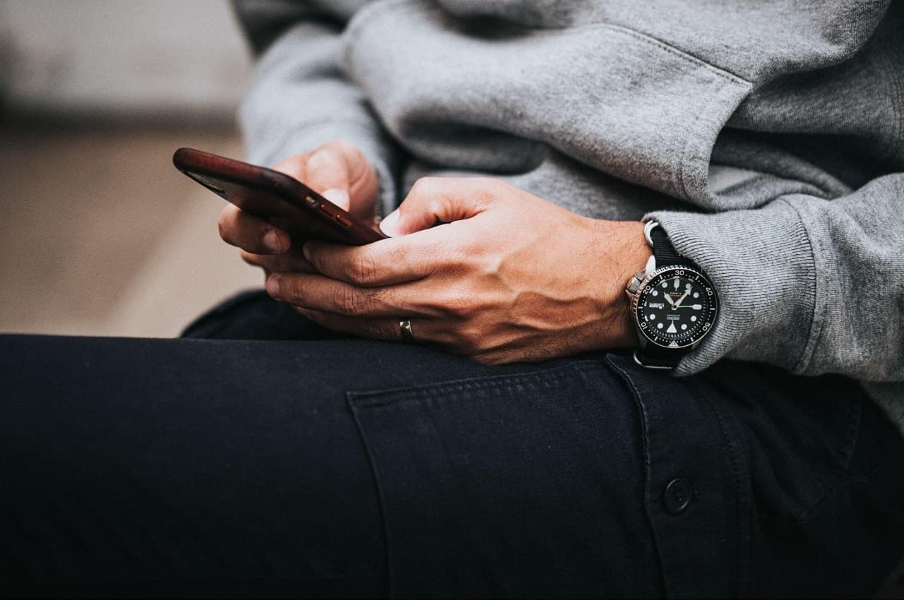 Homem vestindo moletom, relógio e digitando em um celular. O rosto da pessoa não aparece na imagem.