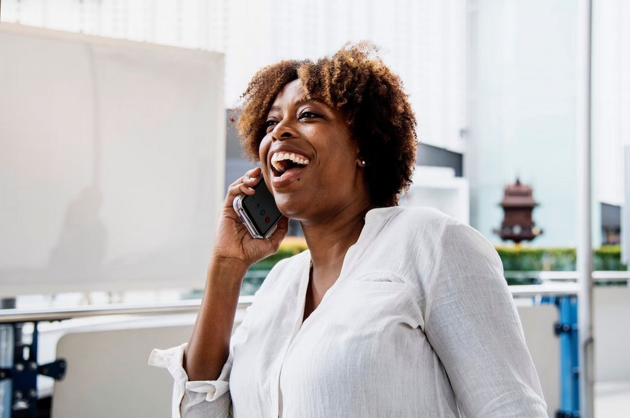 Mulher sorridente ao celular conversando. Ela veste camisa social branca e está no escritório de trabalho.