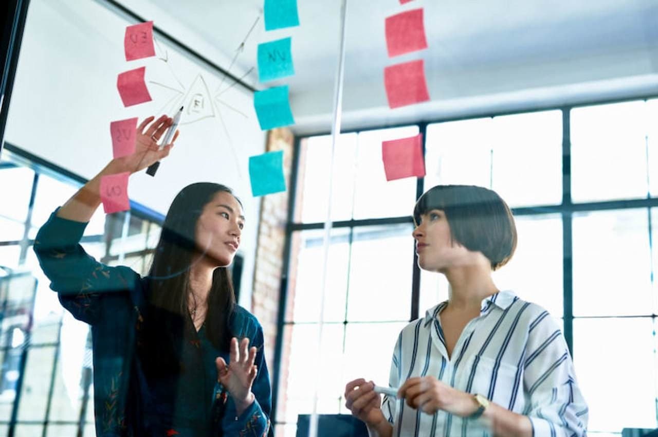 Duas mulheres conversando no ambiente de trabalho e apontando para quadro transparente com post-it rosa e azul colado no quadro.