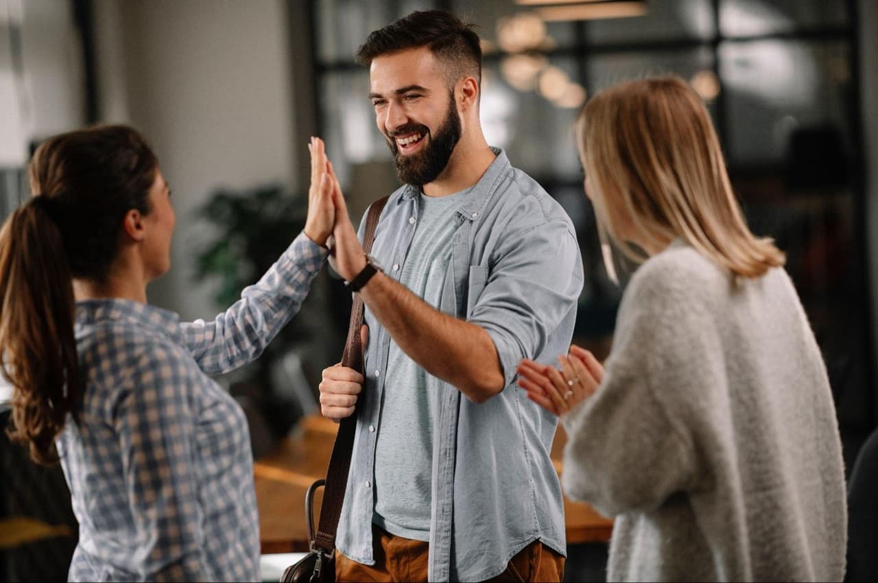 Grupo de pessoas na empresa batendo palmas e se cumprimentando. Na imagem, tem duas mulheres e um homem que segura uma mochila.