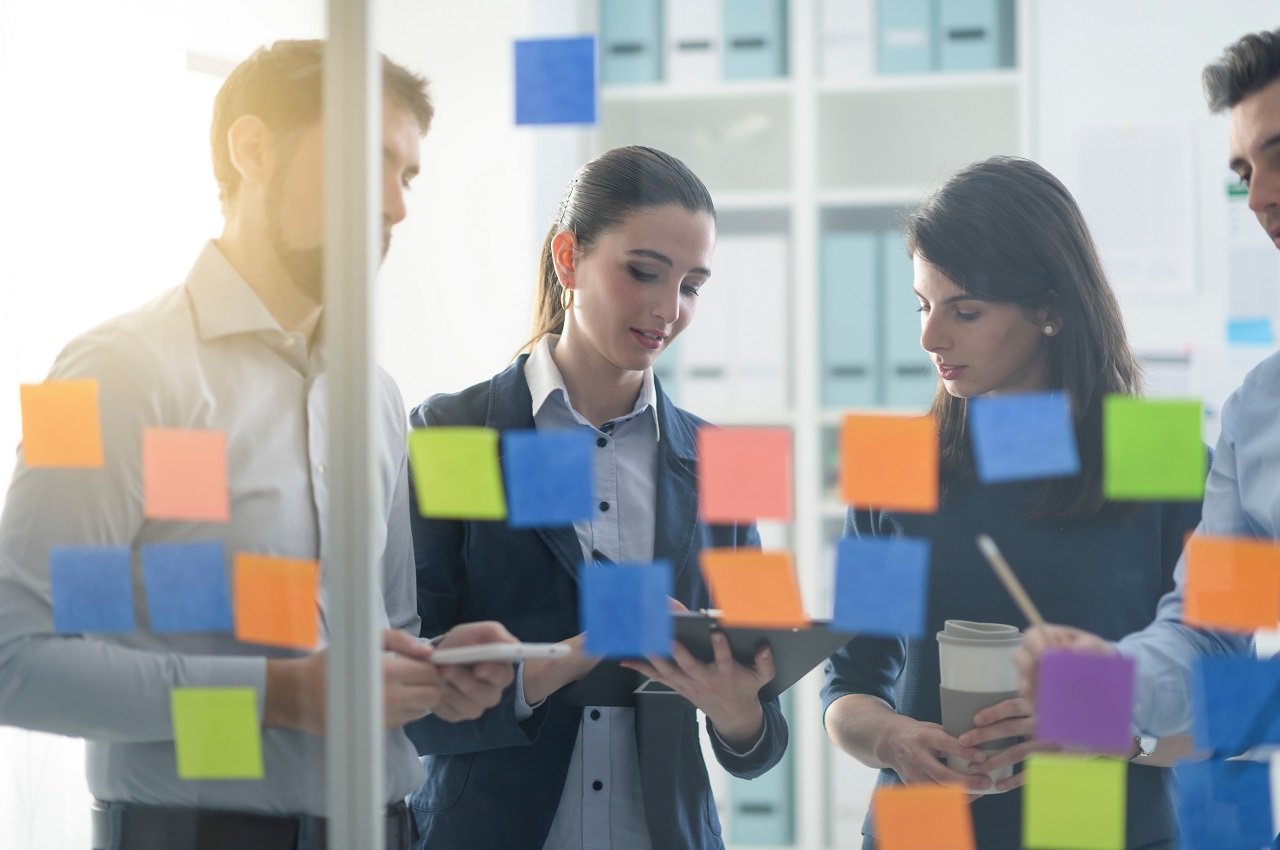 Duas mulheres e um homem realizando anotações em planilhas com expressões concentradas. Na frente do grupo de pessoas, tem um quadro de vidro com post-it coloridas colados.