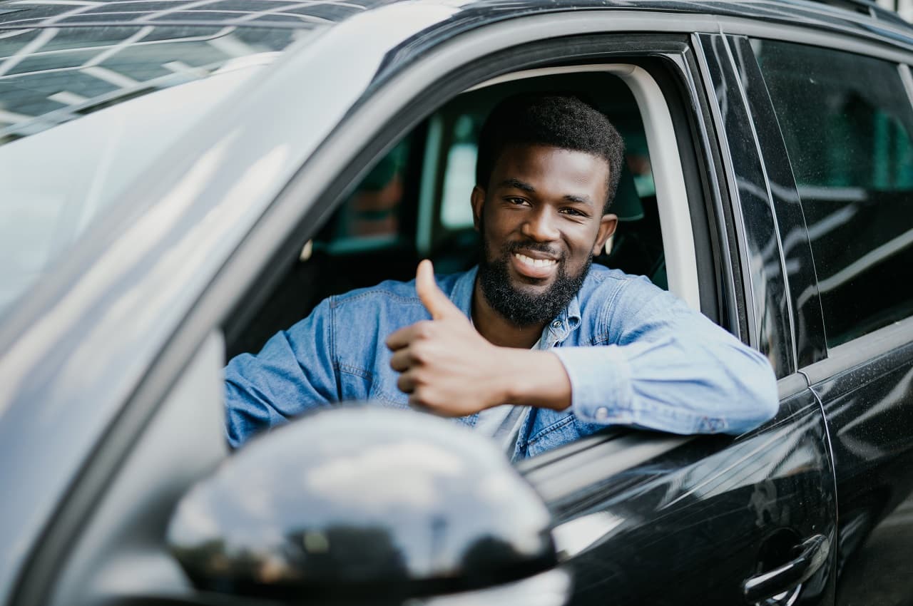 Homem sorridente dirige veículo e apoia o braço esquerdo na lateral do carro, fazendo um joia com a mão.