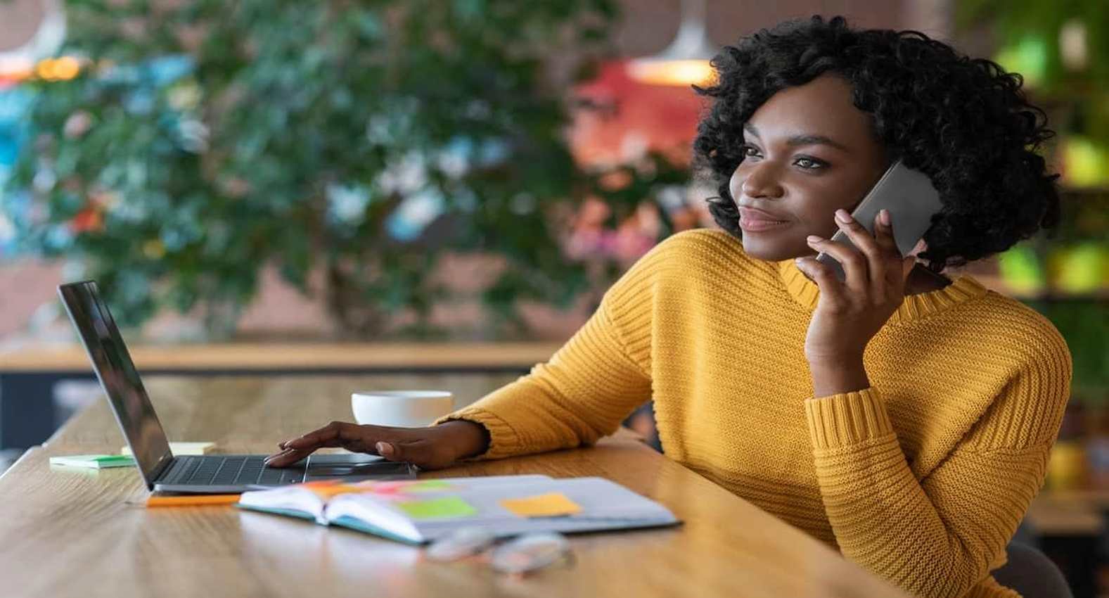 A imagem mostra uma mulher, falando ao telefone e usando seu laptop.