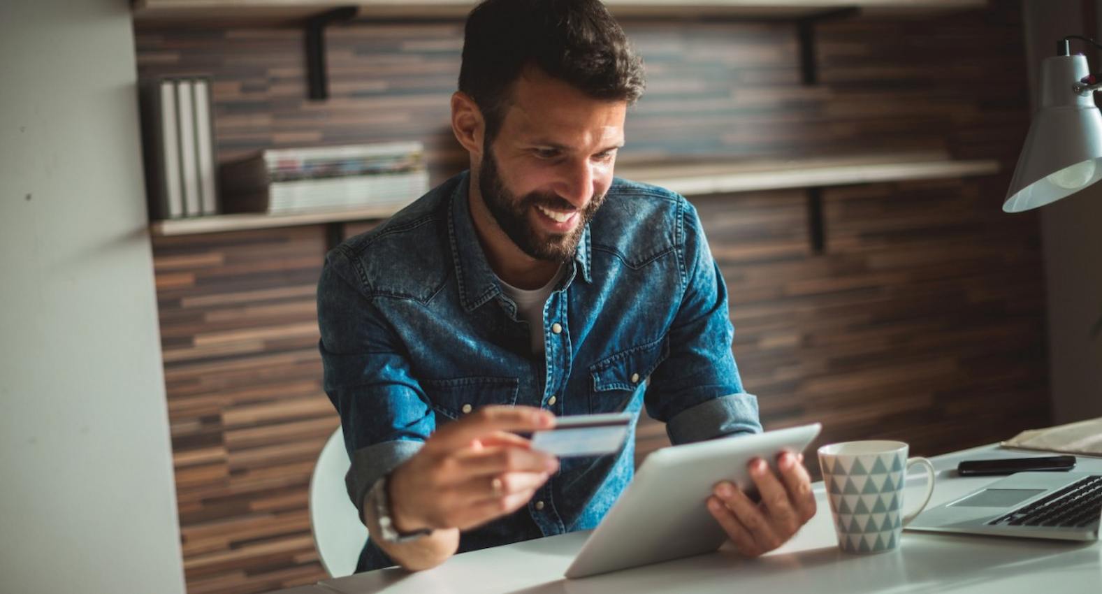 A imagem mostra um homem usando seu tablet e seu cartão de crédito para realizar uma compra. Ao seu lado, uma caneca e um laptop. 