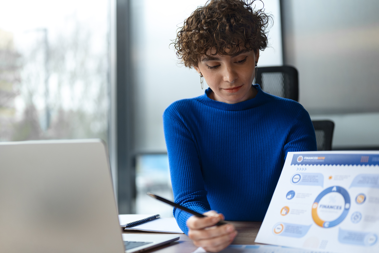 A imagem mostra uma mulher usando uma blusa de mangas longas azul. Ela está usando um laptop e fazendo algumas anotações.