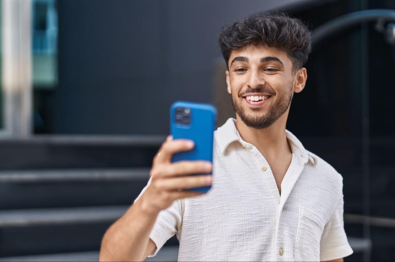 Homem de cabelos castanhos curtos, vestindo camisa de linho branco e sorridente. Ele usa um celular azul escuro para fazer PIX.