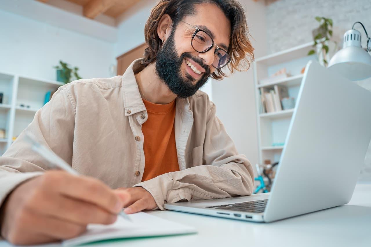 A imagem mostra um homem usando seu laptop e anotando algumas coisas em uma caderneta. Ele está sorrindo para a tela do computador.