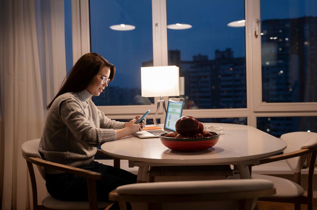 Mulher com expressão concentrada trabalhando a noite através do celular e laptop em cima de sua mesa da cozinha. Ao lado dela, tem um abajur ligado para iluminar seu espaço de trabalho.