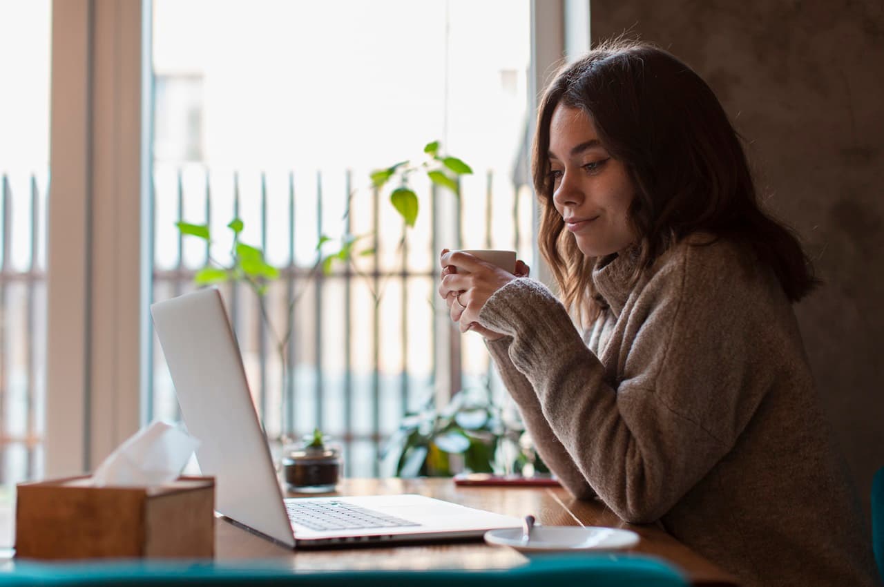 Mulher com expressão pensativa em home office observando tela de laptop da empresa e tomando uma xícara de café. Ela veste suéter bege e está sentada em sua mesa do escritório.