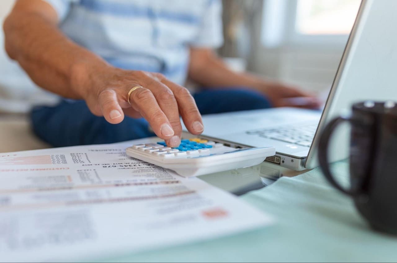 Homem com aliança de casamento dourada no dedo, usando calculadora simples para analisar capacidade produtiva da empresa. Ele usa computador e seu rosto não aparece na imagem.