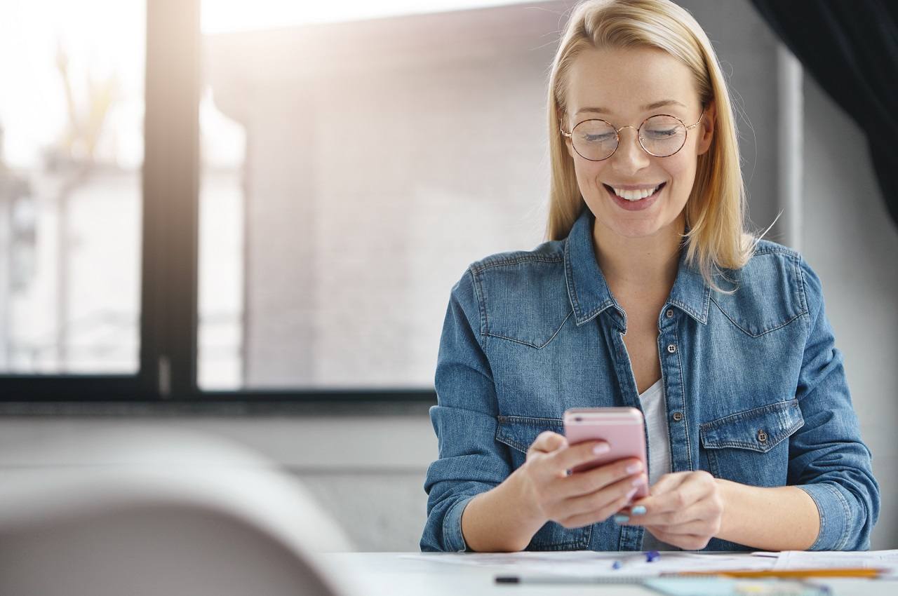 Mulher sorridente de cabelos médios loiros, vestindo camisa branca e jaqueta jeans e usando celular no escritório do trabalho.