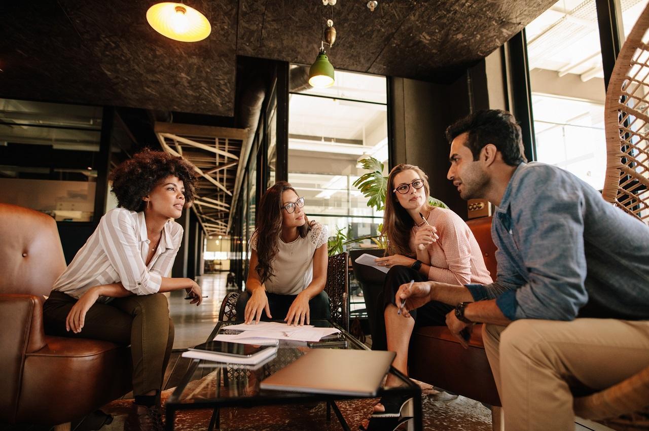 Grupo de colegas de trabalho sentados em sofás e poltronas ao redor de mesa de centro e conversando entre si. Na imagem, tem três mulheres e um homem e todos vestem roupas sociais.