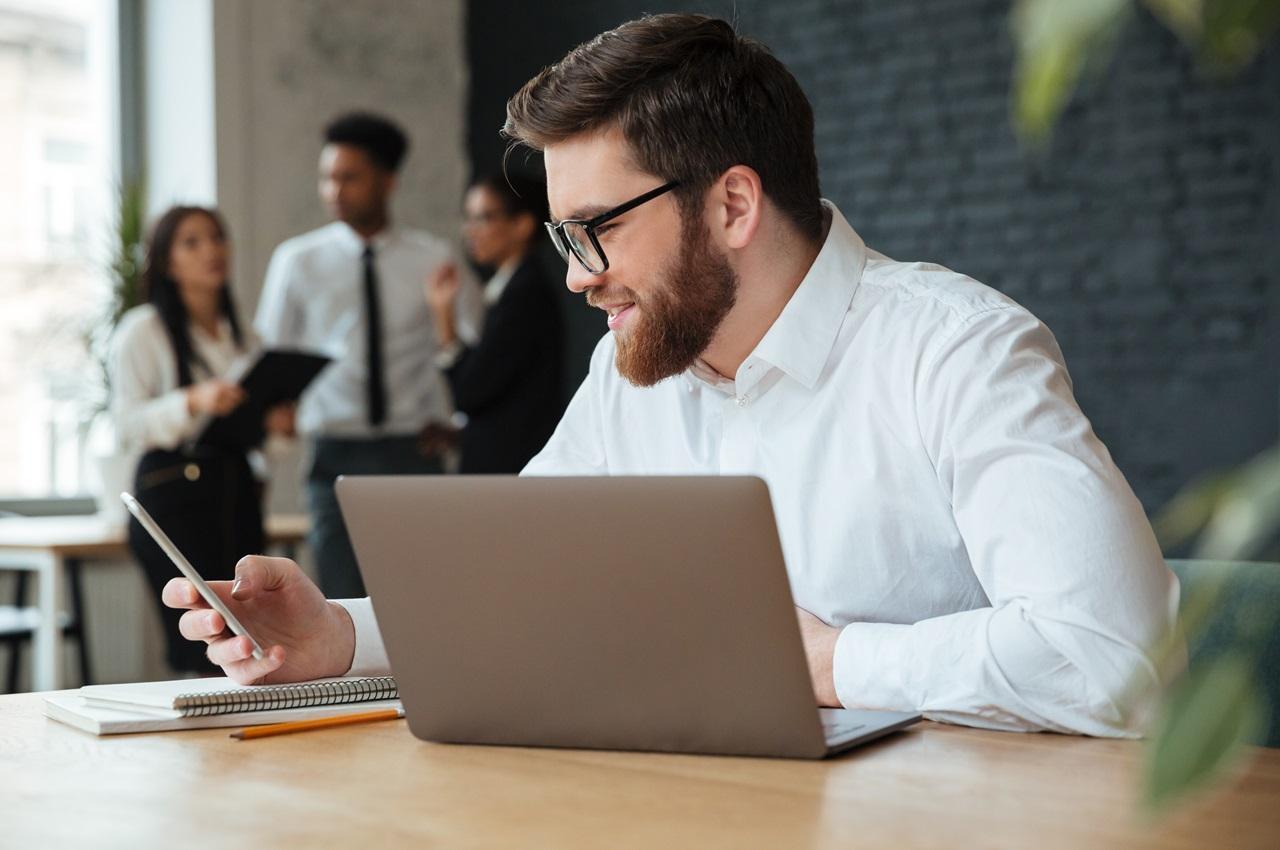 Homem sorridente de camisa social branca e óculos de grau usa um celular e laptop. Atrás do homem, tem um grupo de colegas de trabalho conversando.