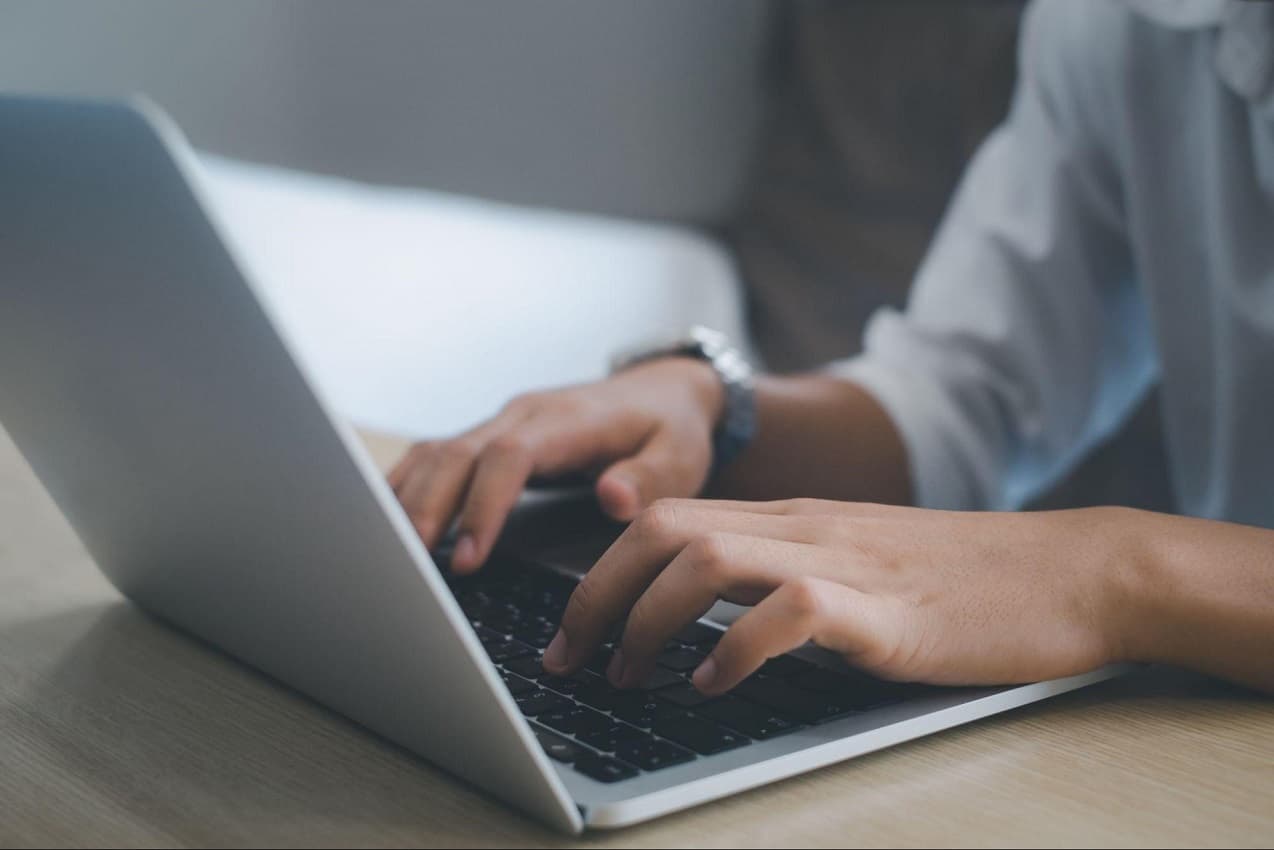 Foco em mãos digitando em teclado de laptop cinza em cima de mesa de madeira no trabalho. O rosto não aparece na imagem e ela veste relógio prata e camisa social azul-clara.