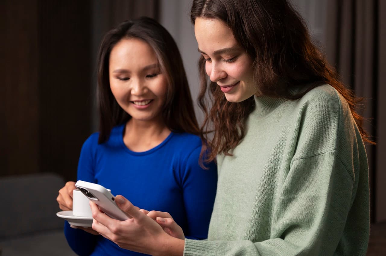 Duas mulheres sorridentes olhando para tela de celular. Uma das mulheres segura xícara de café e veste camiseta azul, enquanto a outra mostra a tela de celular e veste suéter verde-claro.