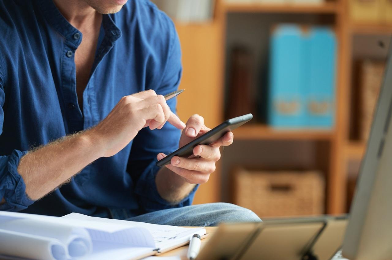 Foco em mão digitando no celular em frente ao laptop e caderno abertos. A pessoa (seu rosto não aparece na imagem) veste blusa social de botões azul escura e está no escritório da empresa.