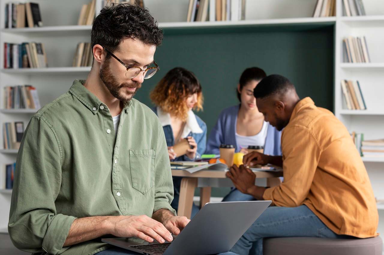 Foco em homem com expressão concentrada que digita no laptop em seu colo. Atrás do homem, tem um grupo de colegas de trabalho que conversam entre eles.