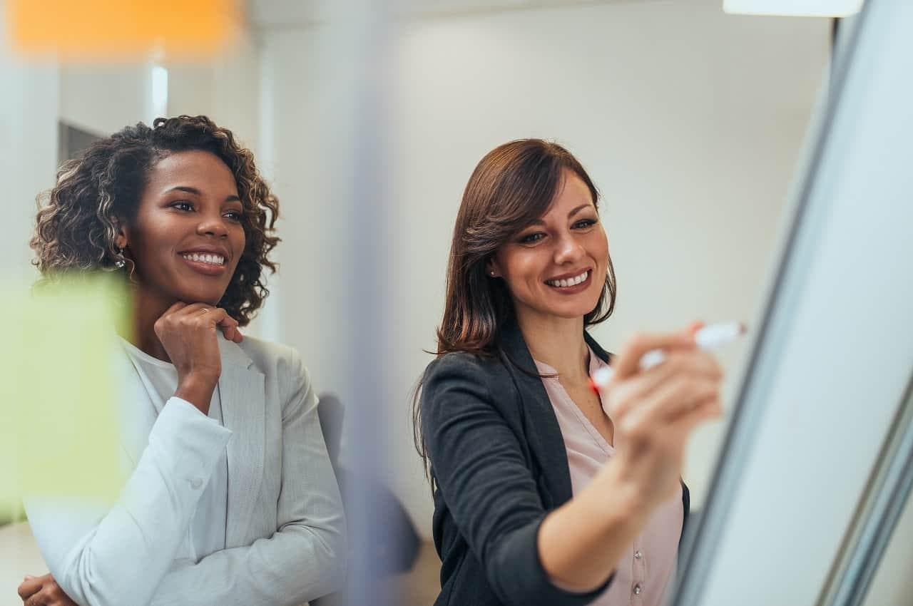 Duas mulheres sorridentes vestindo ternos. Elas usam caneta vermelha e quadro branco para anotar tecnologias no RH.