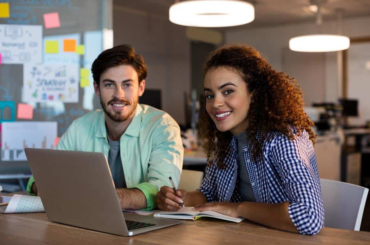 Mulher e homem sorridentes sentados em mesa de escritório e utilizando computador para treinamentos à distância da empresa. A mulher realiza anotações no caderno e o homem segura um gráfico.