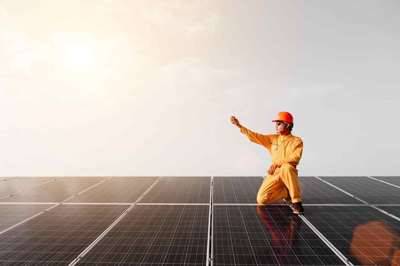 Homem com macacão, capacete e luvas de segurança para energia solar agachado em cima de painéis solares e apontando para o Sol.