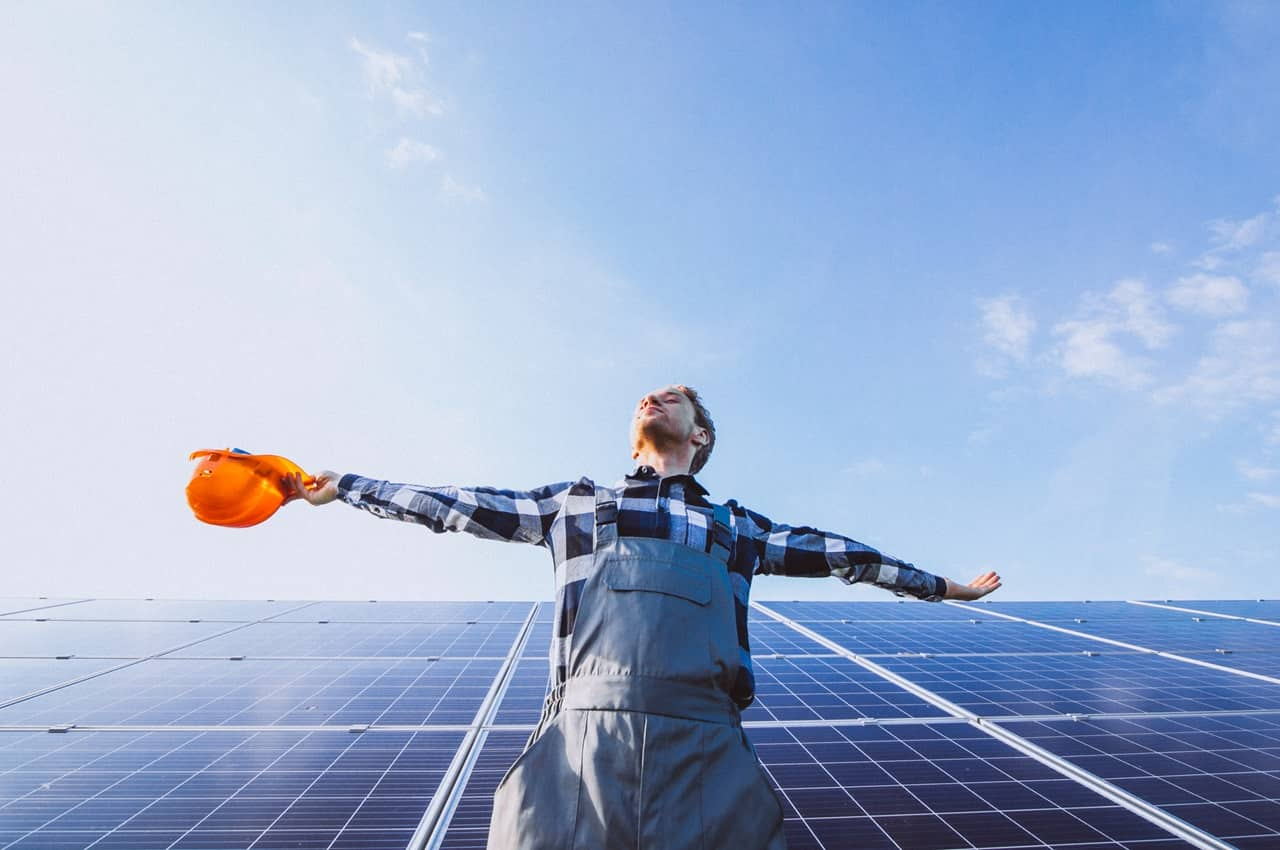 Homem de olhos fechados e braços abertos em frente a uma instalação de geração distribuída de energia solar. O homem veste macacão e capacete de segurança para energia solar.