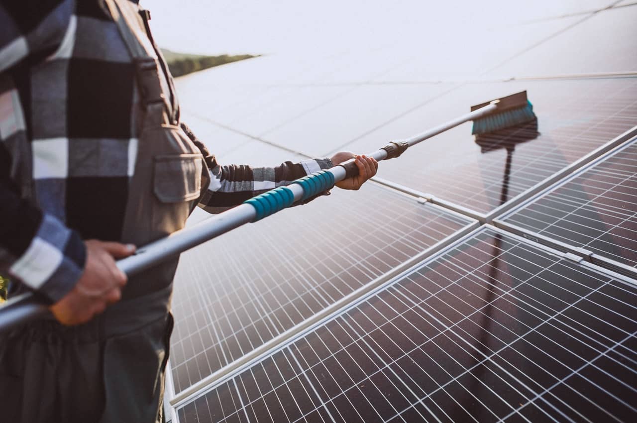 Homem segurando escovão específico para limpeza de placas solares e limpando os painéis instalados. Ele veste macacão de segurança.