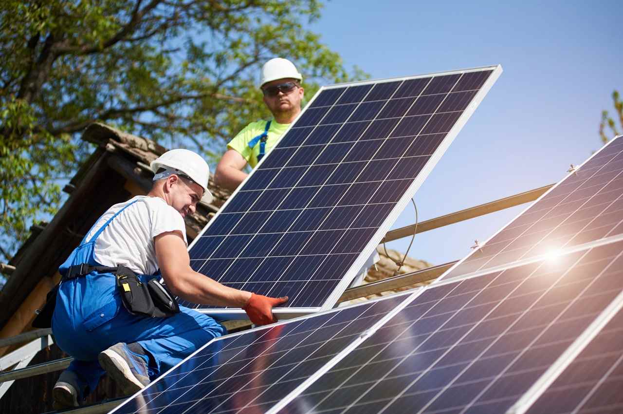 Dois homens sorridentes vestem macacão de proteção, capacete e luvas de proteção para instalação de placas solares. Um dos homens está agachado e faz a manutenção de placas solares.