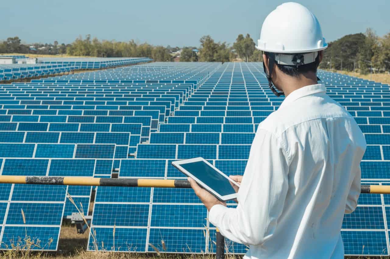 Homem de costas para a imagem veste blusa social branca, capacete de proteção branco e segura tablet nas mãos, enquanto observa placas solares instaladas em plantação.