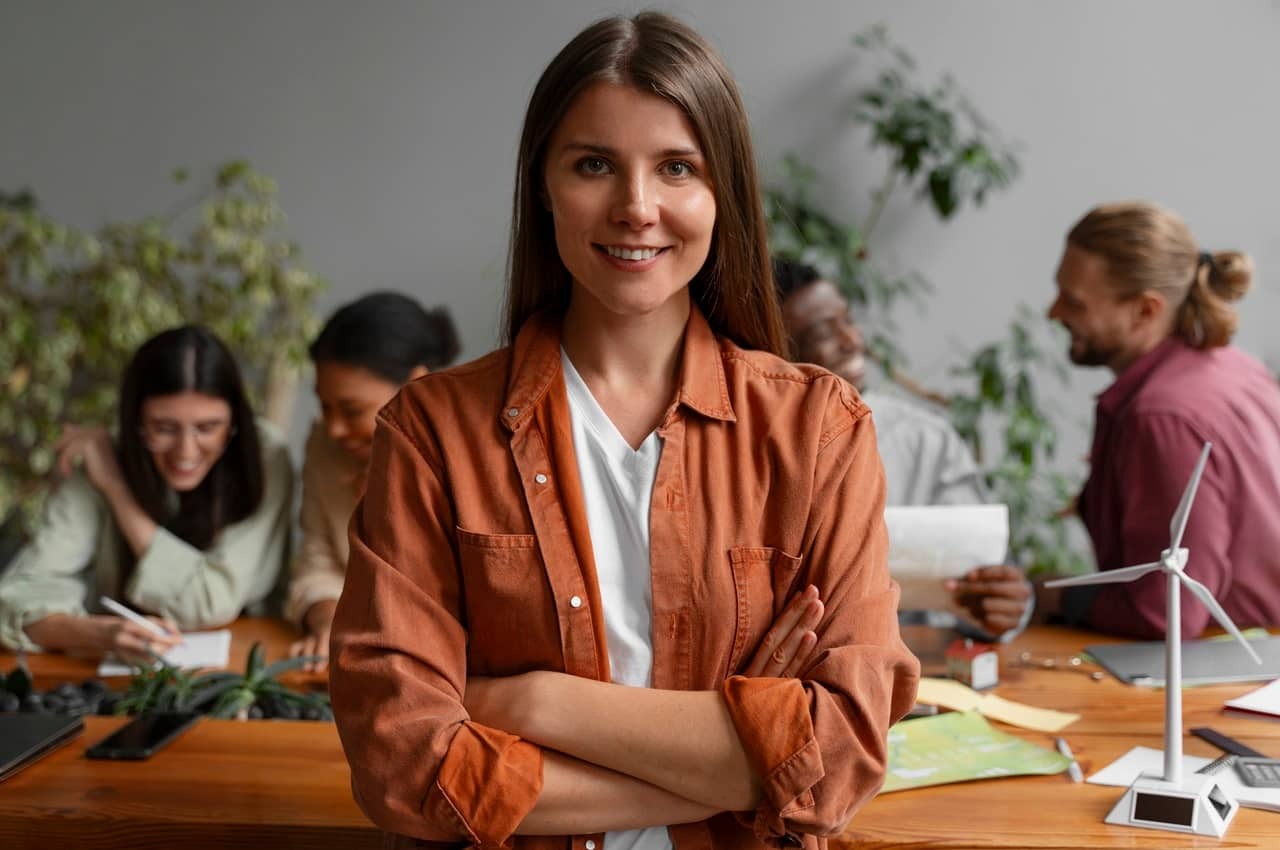 Mulher sorridente no setor de renovável da empresa, em frente a uma mesa de produção inovadora de energia limpa. A mulher veste camisa branca básica e jaqueta terracota por cima e atrás dela tem um time de mulheres trabalhando.