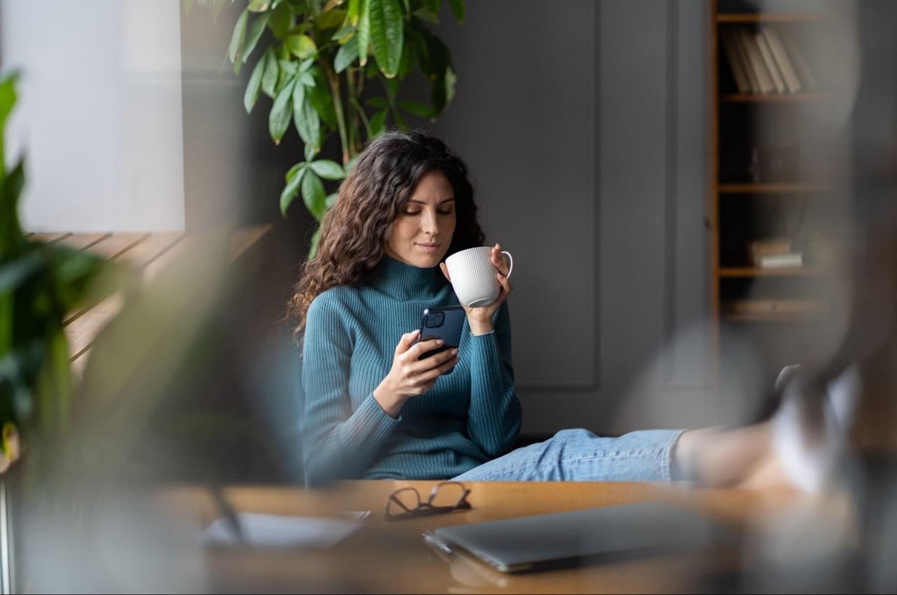 Mulher com expressão tranquila bebe xícara de café e usa celular para saber preços dos painéis solares. Ela coloca os pés em cima da mesa do seu escritório, veste calça jeans e blusa gola alta verde.