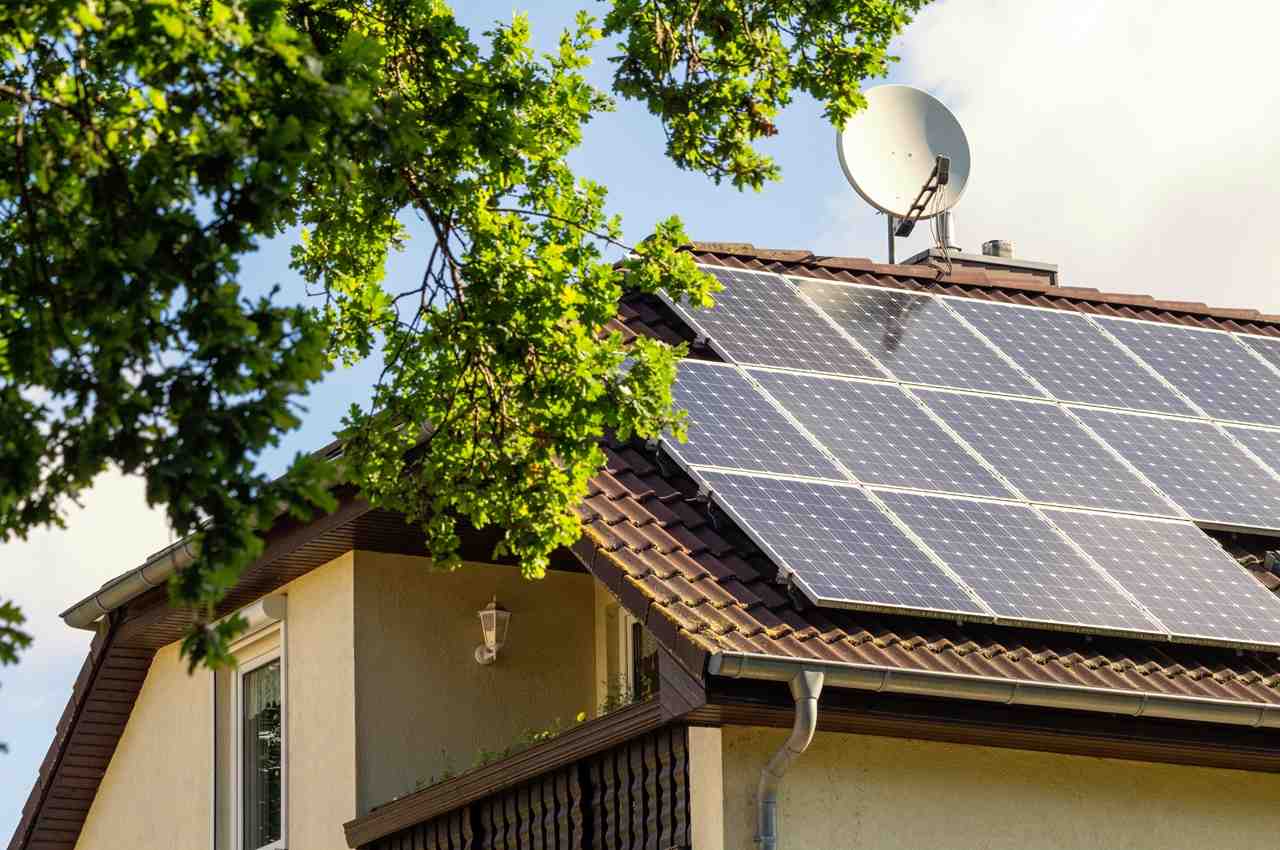 Placas de energia solar instaladas no telhados de uma casa (sobrado) amarela. Em frente a residência, tem uma árvore.