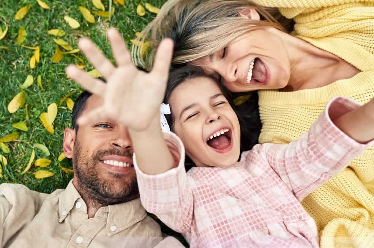 Criança sorridente brincando com duas pessoas adultas (mãe e pai) no gramado de um parque. A mulher veste suéter amarelo, o homem blusa polo bege e a criança vestido rosa de mangas compridas.