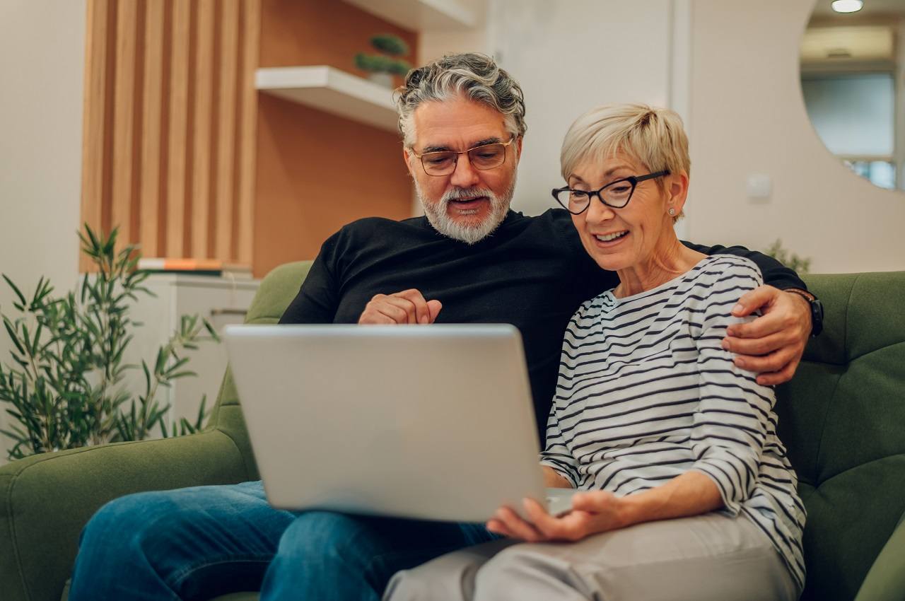 Casal sênior sorridente sentado no sofá usa laptop para anunciar carros na internet. O homem e a mulher usam óculos de grau e estão abraçados. O homem veste suéter azul e a mulher camisa de manga longa listrado.