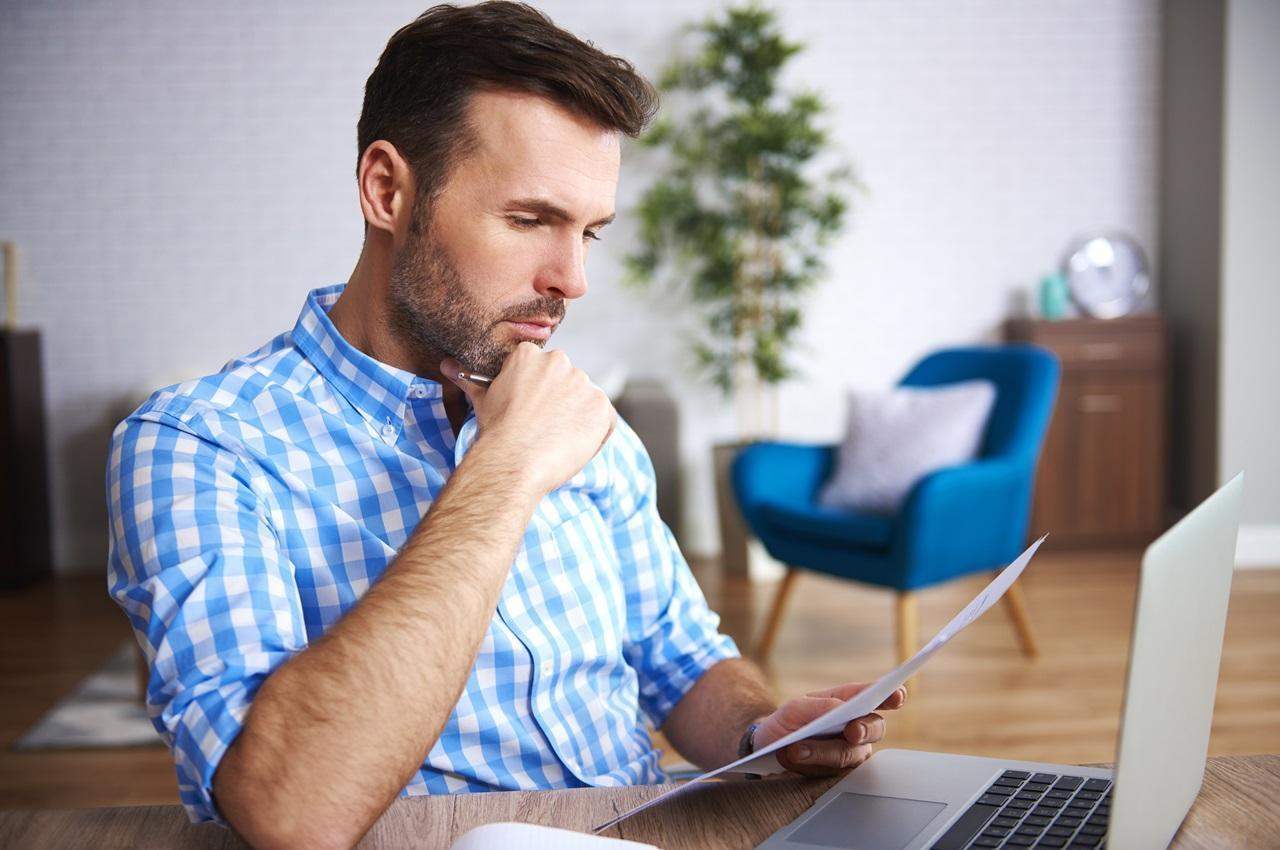 Homem com expressão concentrada apoia a mão no queixo e avalia como calcular a depreciação de veículo pelo laptop. O homem tem cabelos castanhos curtos, barba curta e veste camisa quadriculada azul clara.