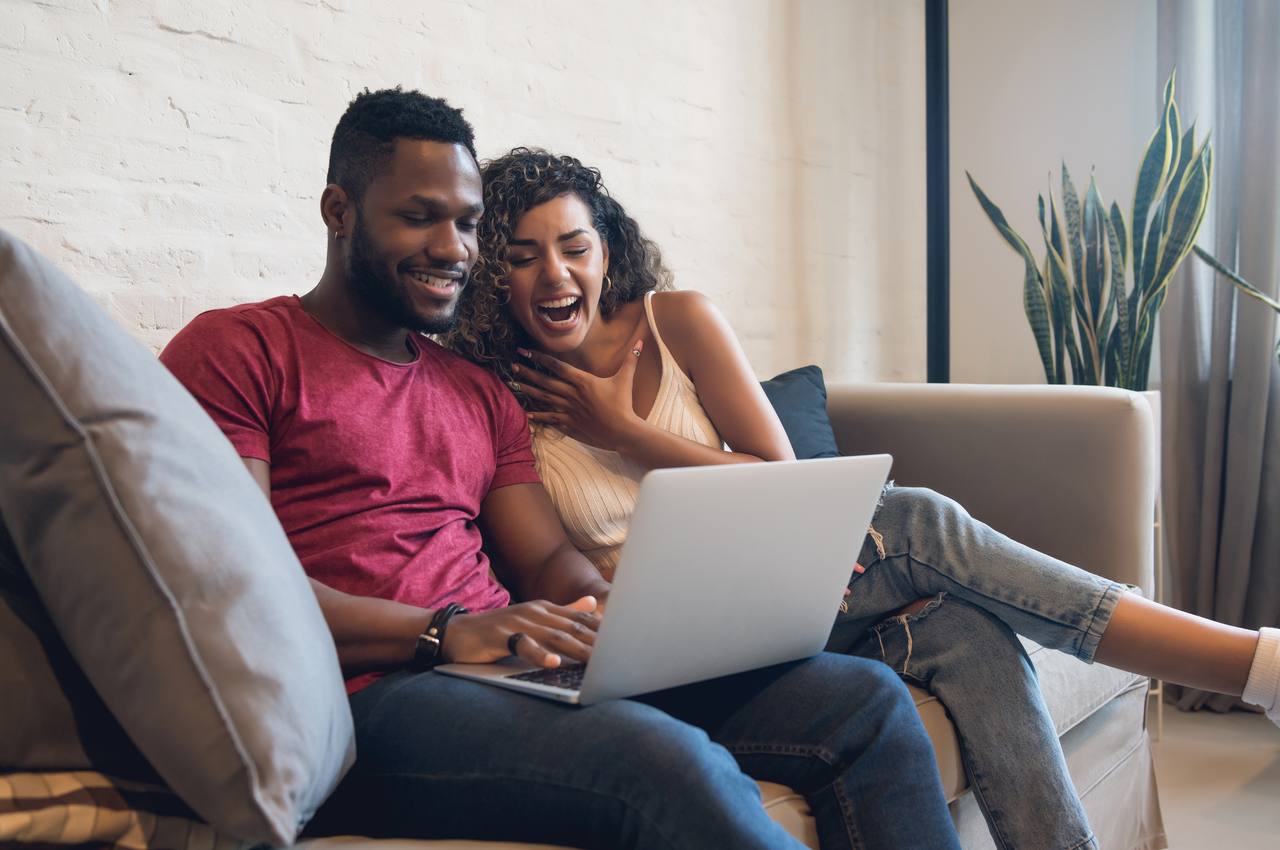 Casal sorridente sentado no sofá de casa usando laptop para comprar carro online. O homem veste blusa vermelha básica e segura laptop no colo, enquanto a mulher se apoia no ombro do homem e sorri.