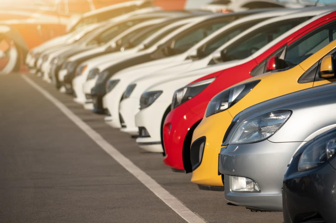 Fileira de carros estacionados de modelos e cores de carros diferentes. Na frente da fila tem carros de cores preta, prata, amarelo, vermelho e branco.