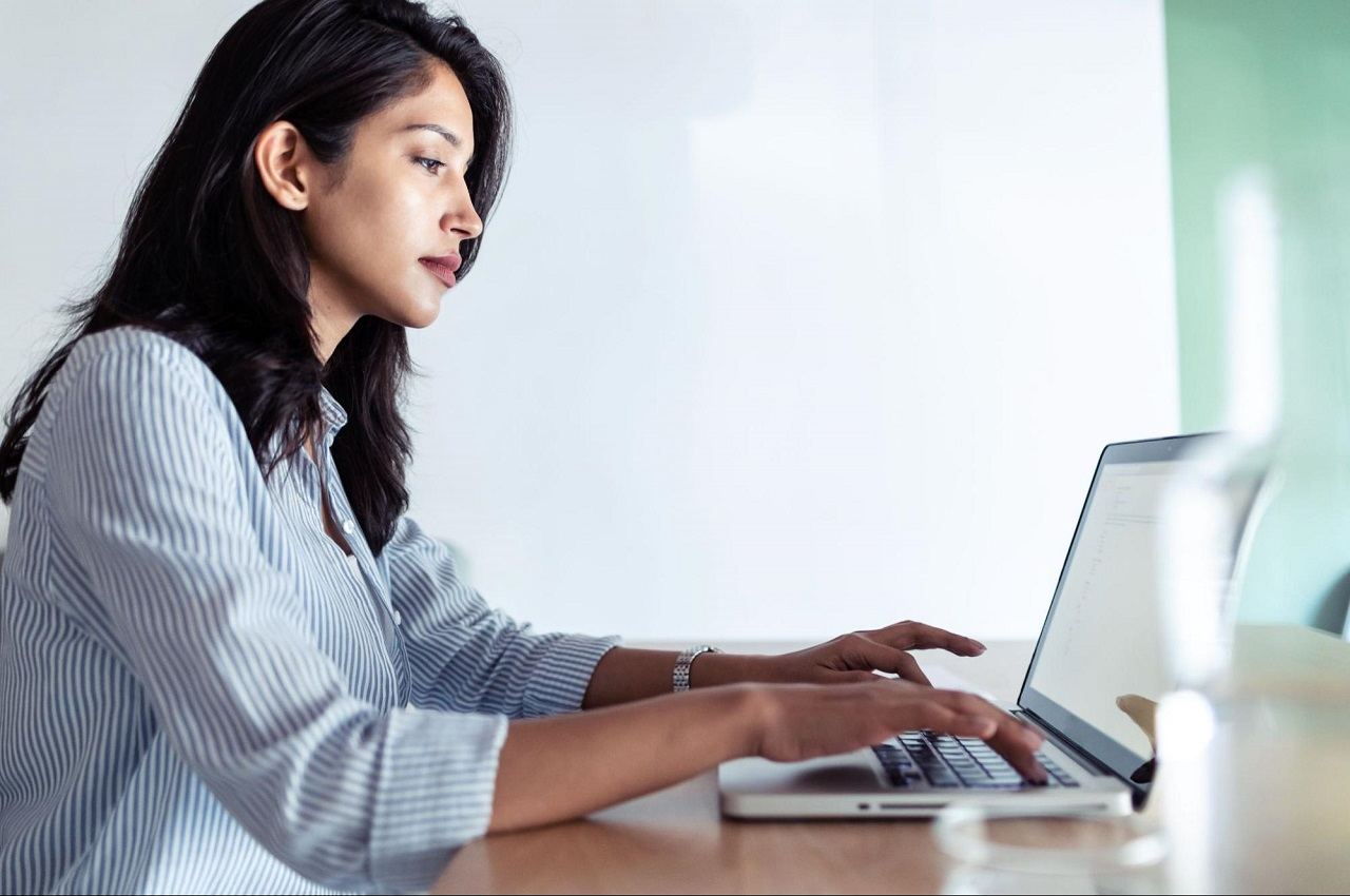 Mulher com expressão concentrada digita no laptop em cima de sua mesa de madeira. A mulher tem cabelos longos pretos e veste blusa social listrada azul e branco.