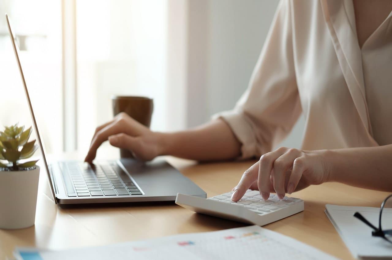 Foco em mão feminina calculando margem de lucro na venda de carro usados pela calculadora e laptop em cima da mesa do seu escritório. O rosto da mulher não aparece na imagem.