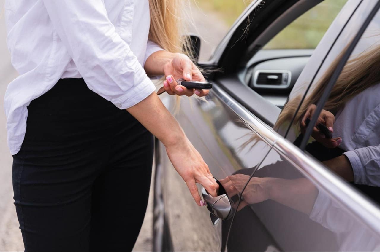 Mulher com roupas sociais abrindo porta de motorista um veículo. Seu rosto não aparece na imagem. 