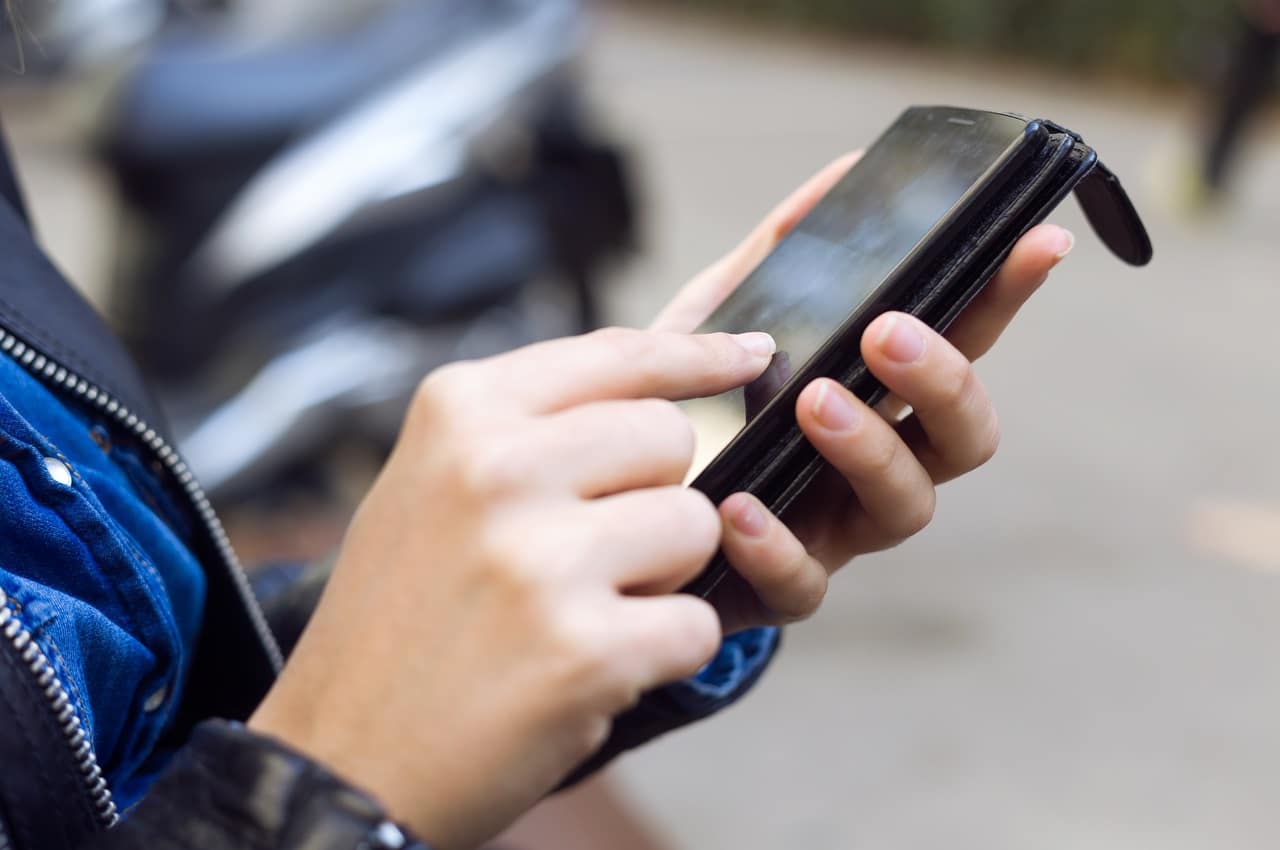 Foco em mulher com dedo indicador tocando na tela de um celular para usar mídias sociais para alavancar os seus negócios. O rosto da mulher não aparece na imagem e ela veste jaqueta de couro preta e blusa jeans.