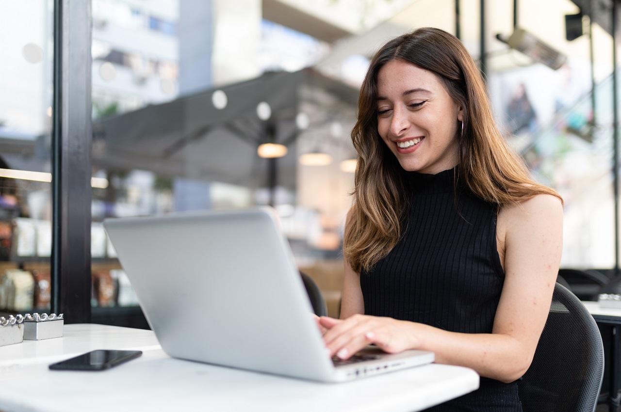 Mulher sorridente de cabelos castanhos longos, sentada na mesa de escritório usando computador para um plano de negócio para loja de automóveis. A mulher veste blusa de gola alta sem mangas na cor preta.