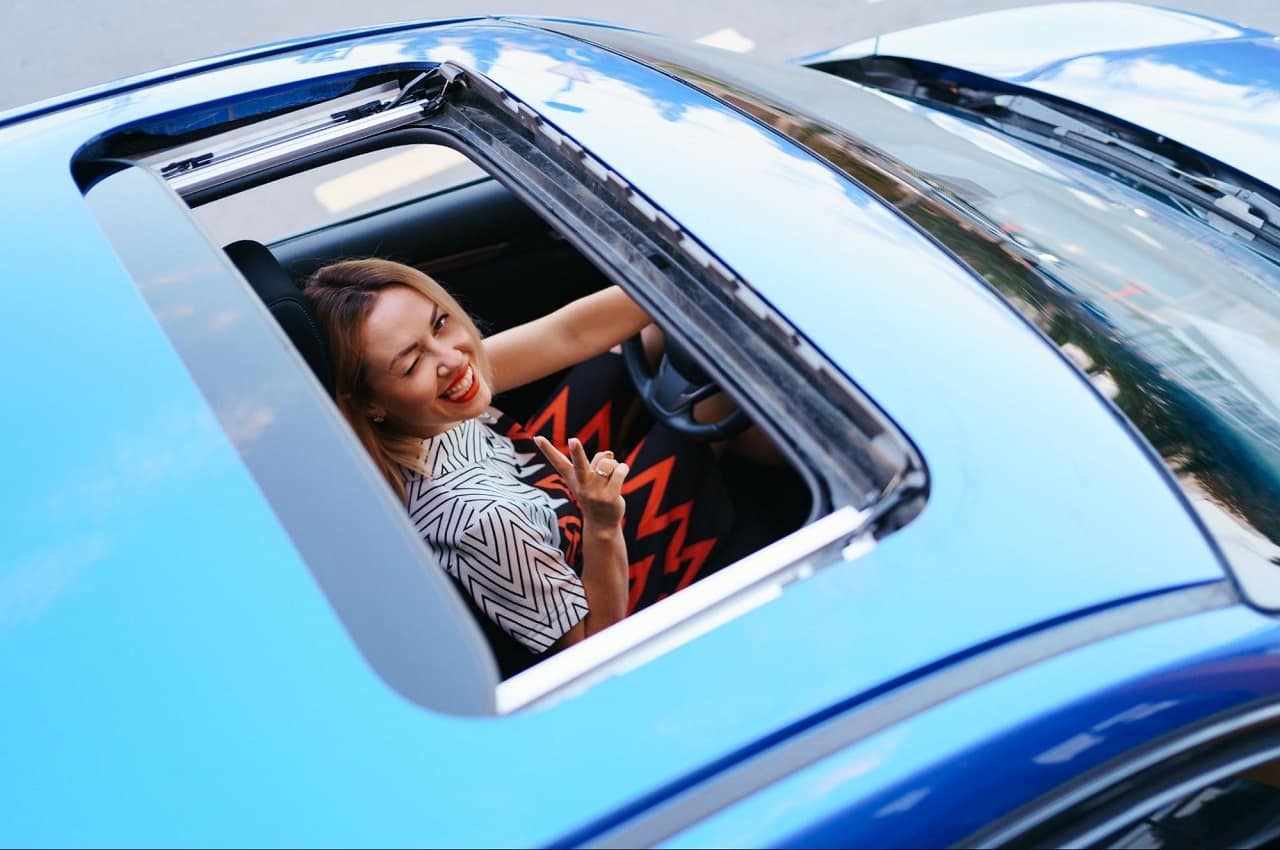 Imagem mostra teto solar de um veículo aberto e uma mulher sorridente no banco de motorista, piscando um dos olhos e olhando pelo teto solar. O veículo é azul claro e a mulher veste um macacão estampado.